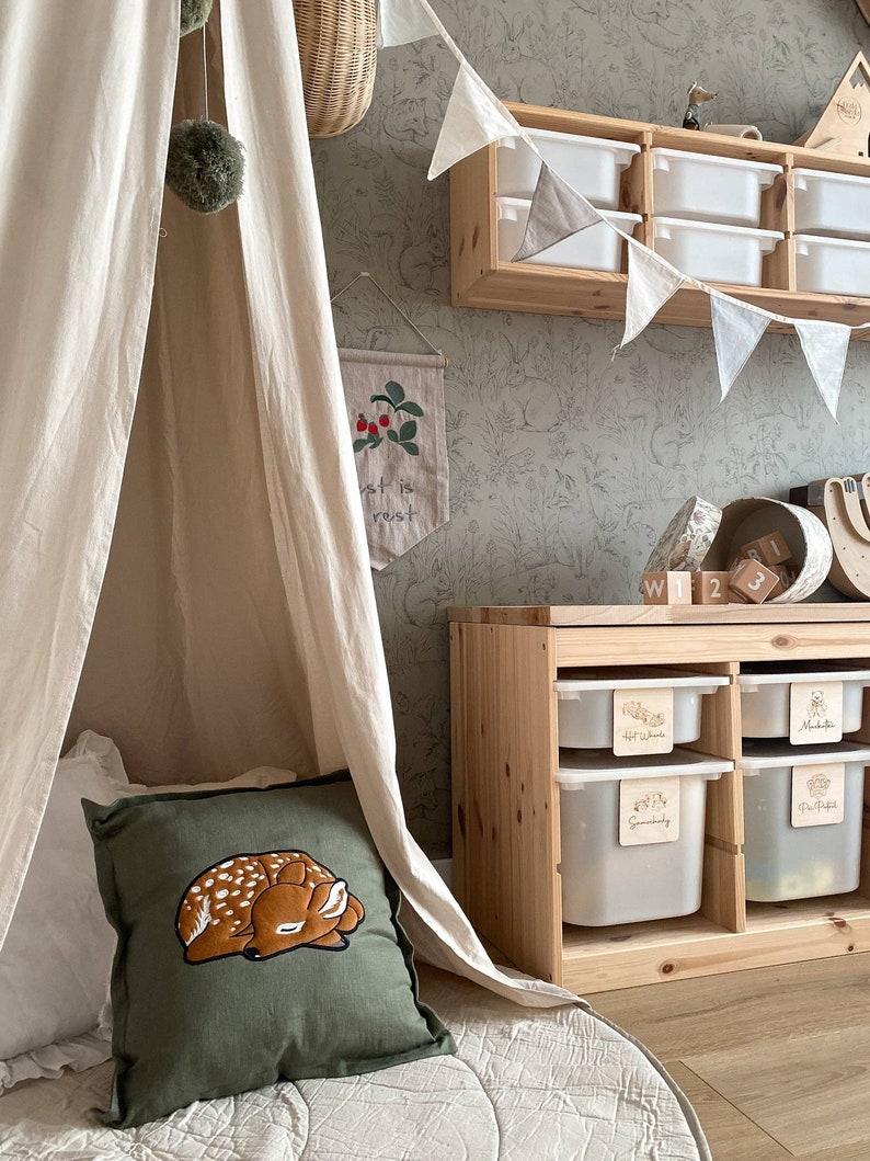 May include: A square olive green throw pillow featuring an embroidered sleeping fawn with white spots. The pillow is positioned inside a cream-colored fabric teepee tent next to a wooden storage unit with white plastic bins and a wall with animal wallpaper.