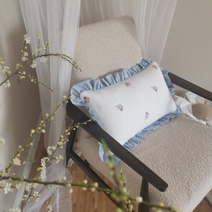 Könnte beinhalten: Ein weißes Kissen mit einem blauen und weißen Gingham-Volant und gestickten Blumen liegt auf einem beigen Sessel mit einem dunklen Holzrahmen. Der Sessel steht vor einem weißen, durchsichtigen Vorhang.