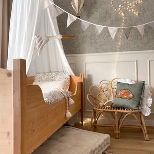 May include: A wooden toddler bed with a white canopy, a floor mattress, and a rattan chair featuring a green pillow with a fawn graphic. The room includes hanging hot air balloon decorations, a string of bunting, and a bird-shaped light fixture.