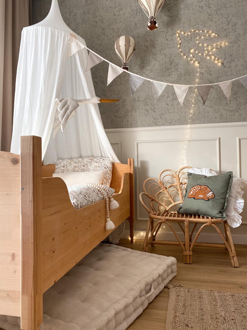 May include: A wooden toddler bed with a white canopy, a floor mattress, and a rattan chair featuring a green pillow with a fawn graphic. The room includes hanging hot air balloon decorations, a string of bunting, and a bird-shaped light fixture.