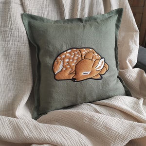 May include: A square olive green throw pillow featuring a central embroidered design of a sleeping fawn. The fawn is brown with white spots and is depicted in a curled, resting position with its eyes closed and ears tucked against its head.