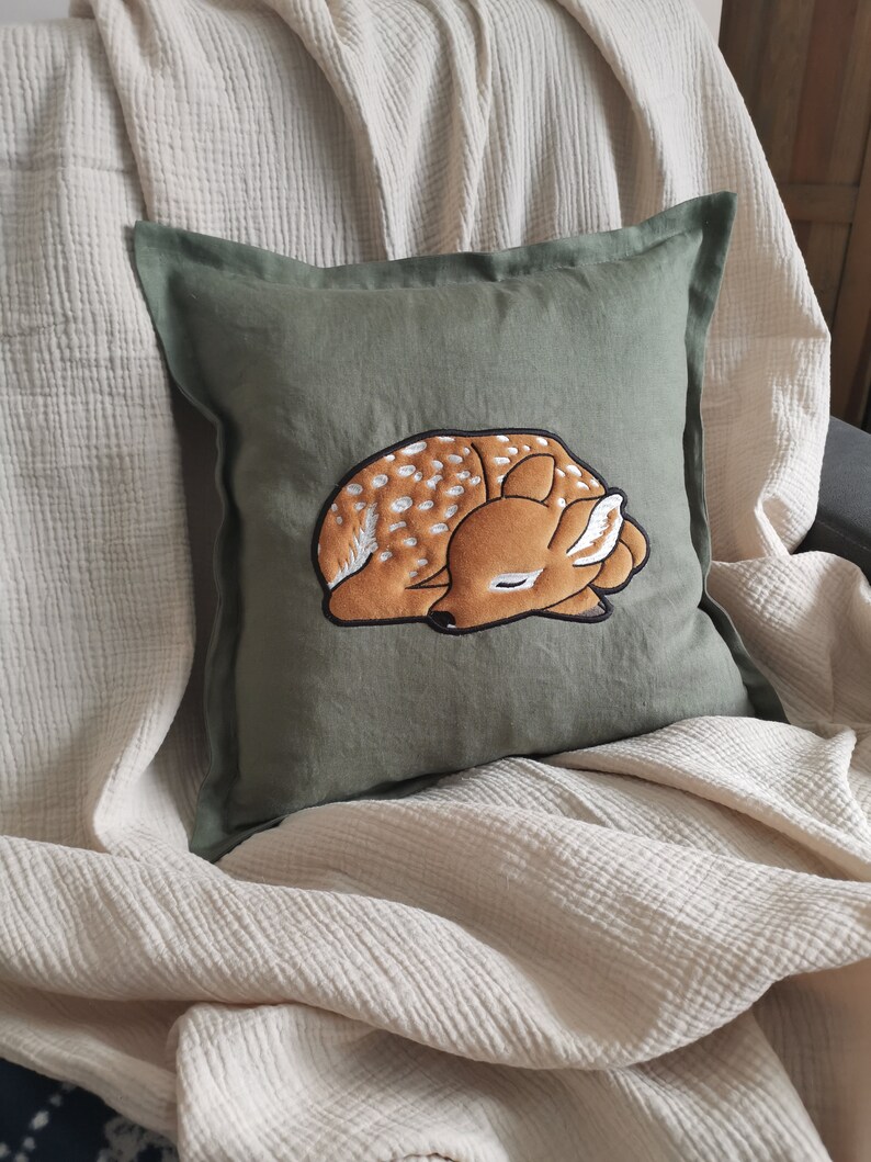 May include: A square olive green throw pillow featuring a central embroidered design of a sleeping fawn. The fawn is brown with white spots and is depicted in a curled, resting position with its eyes closed and ears tucked against its head.