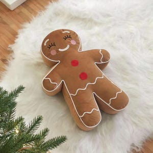 Puede incluir: Un cojín marrón con forma de hombre de jengibre con ribetes blancos y detalles de botones rojos. El cojín tiene los ojos cerrados, mejillas rosas y un contorno blanco. Se coloca sobre una alfombra blanca y esponjosa, con una ramita de hoja perenne en la esquina.