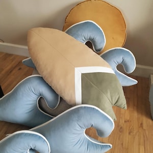 Può includere: Una collezione di cuscini decorativi a tema spiaggia. I cuscini includono una forma di tavola da surf, forme di onde e una forma rotonda di sole. Il cuscino della tavola da surf è beige con un accento bianco e verde. I cuscini a forma di onda sono azzurri con bordi bianchi.