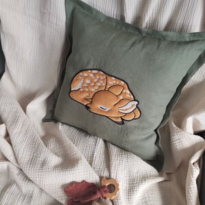 May include: A square olive green pillow featuring a central embroidered graphic of a sleeping fawn with white spots. The fawn is rendered in shades of tan and brown with a dark outline. The pillow has a simple fabric border and a smooth texture.