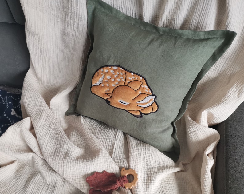 May include: A square olive green pillow featuring a central embroidered graphic of a sleeping fawn with white spots. The fawn is rendered in shades of tan and brown with a dark outline. The pillow has a simple fabric border and a smooth texture.