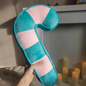 May include: A plush candy cane-shaped pillow in shades of teal and pink. The pillow has a curved shape with alternating color blocks and a teal border. The soft, decorative pillow is held by a hand.