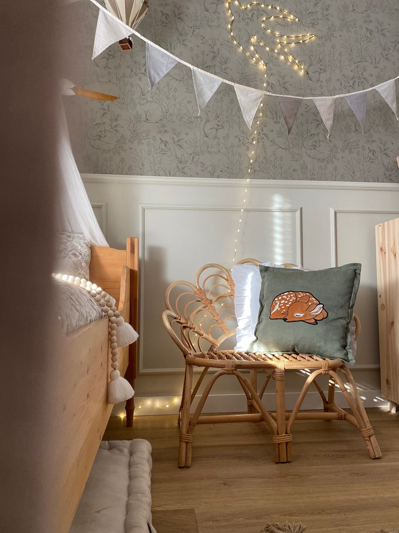 May include: A square olive green throw pillow featuring an embroidered illustration of a sleeping fawn with white spots. The pillow is placed on a light-colored rattan chair with a curved, fan-shaped backrest and four sturdy legs.