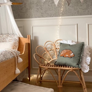 May include: A square olive green throw pillow featuring an embroidered illustration of a sleeping fawn with white spots. The pillow is placed on a light brown curved rattan chair with a fan-shaped backrest, positioned next to a wooden bed frame.