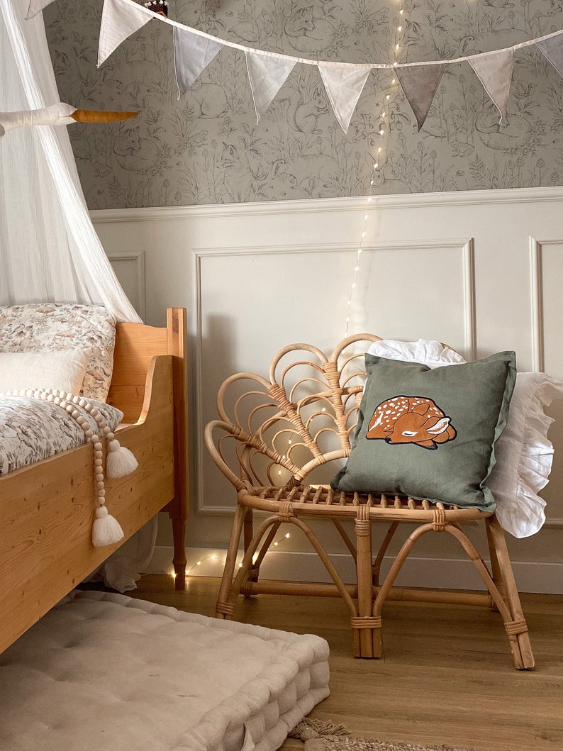 May include: A square olive green throw pillow featuring an embroidered illustration of a sleeping fawn with white spots. The pillow is placed on a light brown curved rattan chair with a fan-shaped backrest, positioned next to a wooden bed frame.