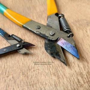 Op de afbeelding: Twee vintage metalen draadknippers met gele en groene handvatten. De grotere draadknipper heeft een veer die aan het handvat is bevestigd. De kleinere draadknipper heeft een blauw handvat.