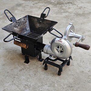 May include: A black metal forge with a square firebox and a hand-cranked blower. The firebox has a wooden handle and a grate. The blower is silver with a wooden handle. The forge is designed for metalworking.