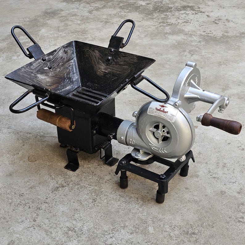 May include: A black metal forge with a square firebox and a hand-cranked blower. The firebox has a wooden handle and a grate. The blower is silver with a wooden handle. The forge is designed for metalworking.