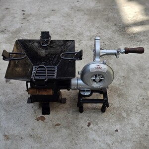 May include: A black metal outdoor stove with a square cooking surface and a hand-cranked blower. The blower is silver with a wooden handle. The stove has a grate and a wooden handle. The stove is designed for outdoor cooking.