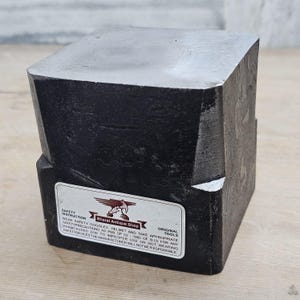 May include: A heavy, black, square-shaped metal anvil block with a smooth, flat top surface. A white rectangular label is affixed to the front, featuring the text Bharat Antique Shop, a hammer logo, and safety instructions regarding protective gear.