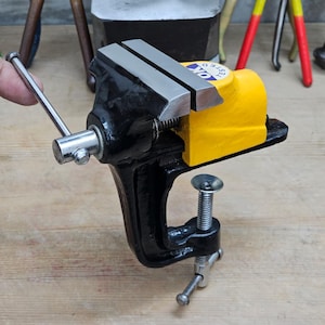 May include: A black and yellow metal bench vise featuring a silver handle and a screw-style clamp base. The top of the yellow housing displays a circular white sticker with the text OK TESTED in blue and black lettering, indicating quality control.