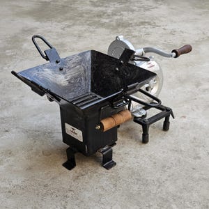 May include: A black metal hand-cranked corn grinder with a square hopper and wooden handles. The grinder has a silver-coloured mechanism and is designed for grinding corn. The grinder is on a concrete surface.