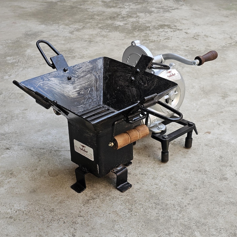 May include: A black metal hand-cranked corn grinder with a square hopper and wooden handles. The grinder has a silver-coloured mechanism and is designed for grinding corn. The grinder is on a concrete surface.