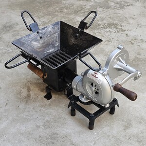 May include: A black metal forge with a square firebox, a grate, and four handles. It has a hand-cranked blower attached, with a silver fan housing and a wooden handle. The forge sits on a concrete surface.