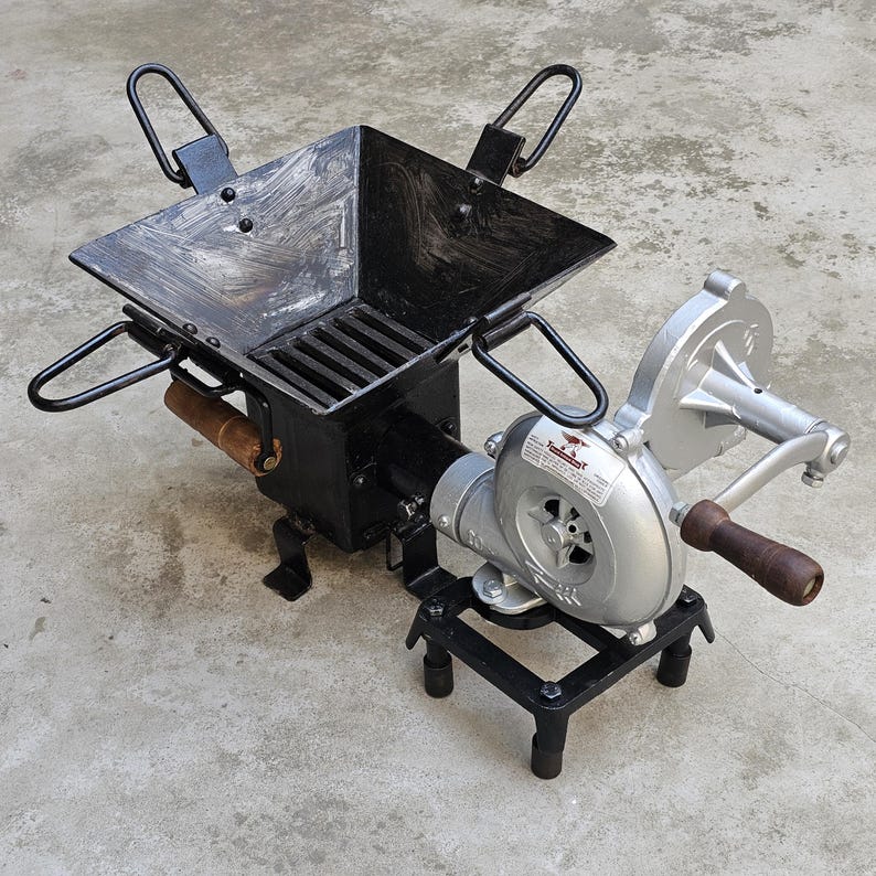 May include: A black metal forge with a square firebox, a grate, and four handles. It has a hand-cranked blower attached, with a silver fan housing and a wooden handle. The forge sits on a concrete surface.