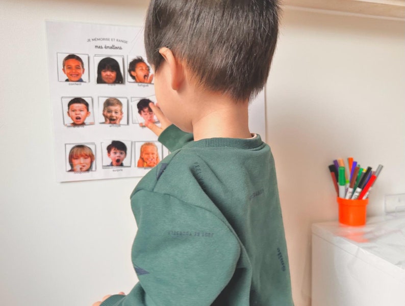 May include: A young child points to a chart with pictures of different facial expressions. The chart is titled "Je memorise et range mes emotions".