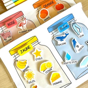 May include: A set of four colourful jars with labels in French. Each jar has a different colour and contains various objects related to that colour. For example, the orange jar contains an orange, a pumpkin, and the word "citrouille" (pumpkin).