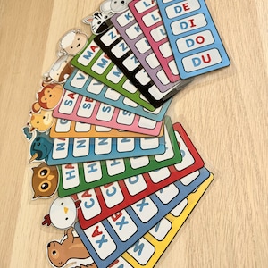 Puede incluir: Un conjunto de tarjetas de memoria coloridas con ilustraciones de animales y letras del alfabeto. Cada tarjeta presenta un animal diferente y una letra, la letra se repite varias veces en la tarjeta. Las tarjetas est&aacute;n dispuestas en forma de abanico, con la tarjeta superior mostrando un delf&iacute;n y las letras DA, DE, DI, DO y DU.