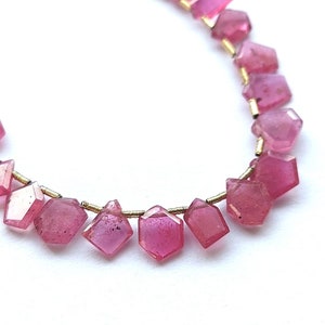 Pode incluir: Um fio de contas de pedras preciosas cor-de-rosa com um fio de ouro enfiado pelo centro de cada conta. As contas são facetadas e têm uma forma ligeiramente irregular.