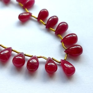 May include: A strand of deep red gemstone beads in a teardrop shape. The beads are strung on a gold wire.