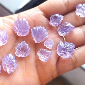 May include: A collection of small, carved lavender-colored gemstone beads in various shapes, including leaves, shells, and flowers. The beads are translucent and appear to be made of amethyst or a similar material. The intricate carvings add detail to each piece.