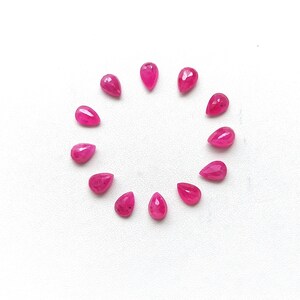 May include: A collection of eleven teardrop-shaped, faceted, bright pink gemstones arranged in a circle on a white surface. The gems have a polished, reflective surface, and vary slightly in size and shape.