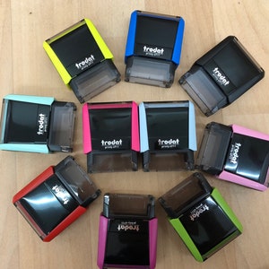 Könnte beinhalten: Eine Sammlung von zehn verschiedenen selbsttintenenden Stempeln in verschiedenen Farben mit dem aufgedruckten Wort "trodat". Die Stempel sind in einem Kreis auf einer Holzoberfläche angeordnet.