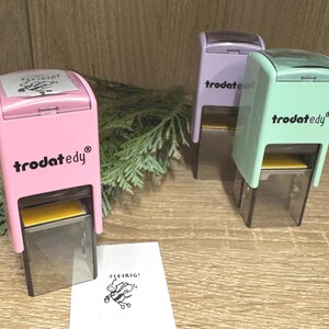 Könnte beinhalten: Drei selbstfärbende Stempel in Rosa, Lavendel und Mintgrün. Jeder Stempel trägt den Markennamen "trodat edy". Ein gedrucktes Bild einer Biene mit dem Wort "Fleibig!" befindet sich auf einem weißen Papier.