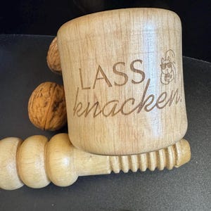 Könnte beinhalten: Ein Nussknacker aus Holz mit einem Eichhörnchen-Design und dem Text "LASS knacken". Der Nussknacker hat einen Griff mit einem Gewinde am Ende.