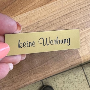 K&ouml;nnte beinhalten: Ein rechteckiges Schild in Goldfarbe mit dem Text "keine Werbung" in schwarzer Schrift.