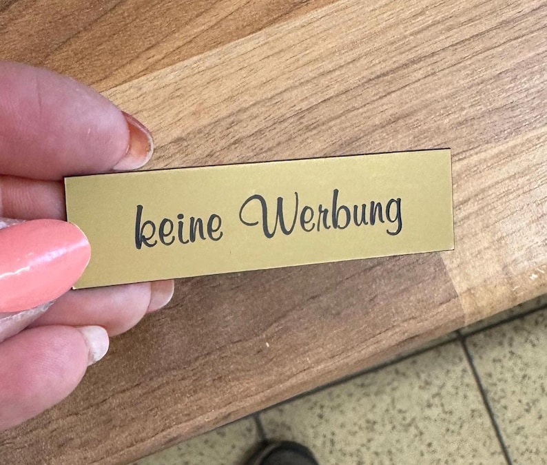 K&ouml;nnte beinhalten: Ein rechteckiges Schild in Goldfarbe mit dem Text "keine Werbung" in schwarzer Schrift.