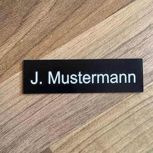 K&ouml;nnte beinhalten: Schwarzes Namensschild mit wei&szlig;em Text "J. Mustermann"