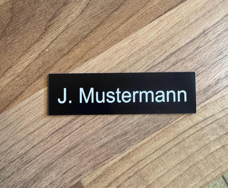 K&ouml;nnte beinhalten: Schwarzes Namensschild mit wei&szlig;em Text "J. Mustermann"