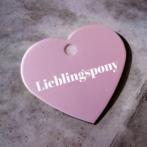 May include: A pink heart-shaped tag with a hole at the top and the text "Lieblingspony" in white.