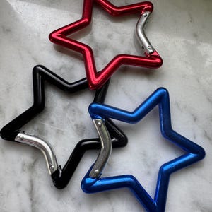 Könnte beinhalten: Drei sternförmige Karabiner in Rot, Schwarz und Blau. Jeder Karabiner verfügt über eine glatte, metallische Oberfläche und einen silberfarbenen, federbelasteten Verschlussmechanismus, der ein sicheres Öffnen und Schließen ermöglicht.