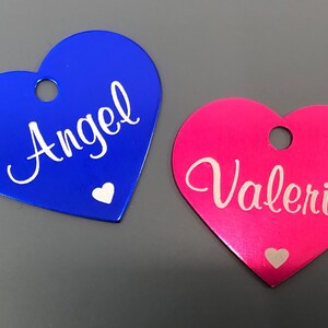May include: Two heart-shaped metal tags with white lettering. The blue tag says "Angel" and the pink tag says "Valeria".