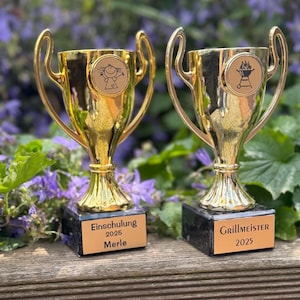 Op de afbeelding: Twee goudkleurige trofeeën met zwarte sokkels. Eén trofee heeft een stokfiguurontwerp, de andere een grillontwerp. Op de trofeeën staan de woorden "Einschulung 2025 Merle" en "Grillmeister 2025".