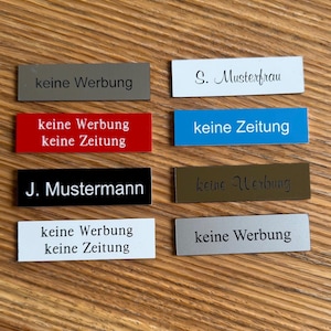 May include: Eight rectangular signs with black text on different colored backgrounds. The signs say "keine Werbung" (no advertising) and "keine Zeitung" (no newspaper). Two signs have names on them: "S. Musterfrau" and "J. Mustermann".