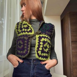 May include: A sleeveless crochet vest constructed from square motifs in shades of lime green, dark purple, and olive green. The vest features a front tie closure with matching yarn strings and a textured, open-work pattern throughout the garment.