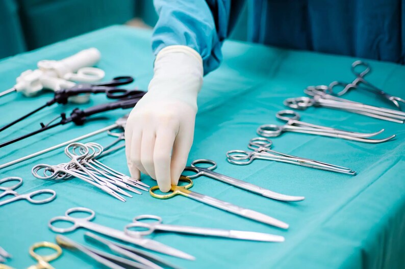 May include: A selection of surgical instruments arranged on a blue surgical drape. The instruments include forceps, scissors, and a needle holder. A gloved hand is reaching for a pair of forceps.