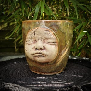 Może przedstawiać: Ceramiczny kubek z wyrzeźbioną twarzą śpiącego dziecka. Kubek jest glazurowany w odcieniach zieleni i brązu, z twarzą w jaśniejszym, kremowym kolorze. Kubek jest cylindryczny i stoi na ciemnej powierzchni.