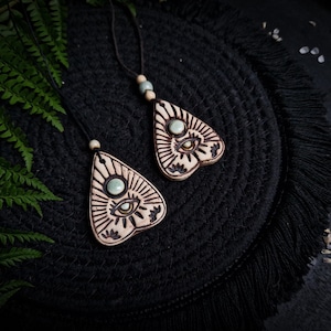 Könnte beinhalten: Zwei Holzanhänger mit Planchette-Design, mit einem Auge und Sonnenstrahlenmuster und einem hellgrünen Stein. Die Anhänger hängen an schwarzen Kordeln mit Holzperlen. Der Hintergrund ist eine dunkle, strukturierte Oberfläche mit Farnblättern.