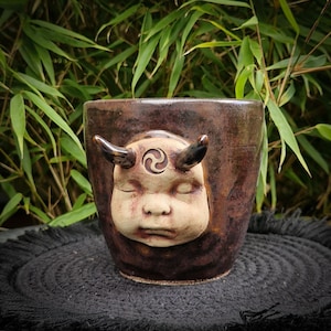 Op de afbeelding: Een bruine keramische plantenpot met een gebeeldhouwd gezicht van een slapende figuur met hoorns en een spiraalsymbool op het voorhoofd. De pot heeft een getextureerd oppervlak en staat tegen groene bladeren.