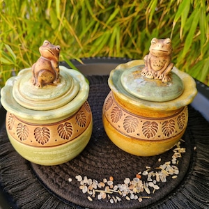 Op de afbeelding: Twee keramische potten met deksels, elk bekroond met een kikkerfiguur. De potten zijn groen en bruin met bladerpatronen. De potten staan op een zwarte, gefranjerde mat, met een groene, bladerrijke achtergrond.