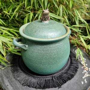 Peut inclure: Un pot en céramique vert moucheté de forme ronde avec deux anses latérales et un couvercle assorti. Une petite figurine de grenouille brune sculptée est placée sur le dessus du couvercle en guise de poignée. Le pot a une finition mate texturée.